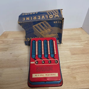 Puede incluir: Una máquina de sumar vintage Wolverine No. 39 con carcasa de metal rojo y ranuras verticales azules que contienen los números del uno al nueve. El dispositivo cuenta con una ventana de visualización total con cuatro ceros y su caja azul.