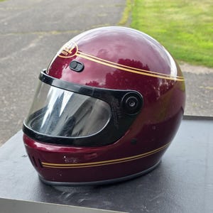 Puede incluir: Un casco de moto integral de color granate oscuro con acabado brillante, que presenta finas rayas doradas que rodean la carcasa. El casco incluye una visera transparente con marco negro, ventilación en la barbilla y el logo de Bell Helmets.