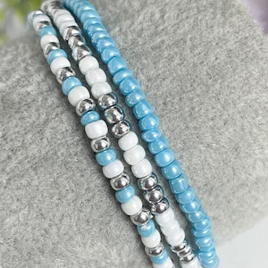 May include: Three beaded bracelets in shades of blue and white. The bracelets are made with small, round beads and are layered on top of each other.