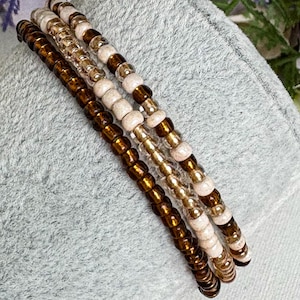 May include: Four beaded bracelets in shades of brown, tan, and gold. The bracelets are made of small, round beads strung together. The beads have a glossy finish and are arranged in a spiral pattern. The bracelets are displayed on a soft, grey surface.