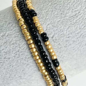 May include: A close-up of a layered bracelet made of black and gold beads. The bracelet is made of multiple strands of beads, with the black beads being smaller than the gold beads.