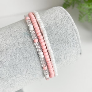 May include: Three layered beaded bracelets in pink and white. The bracelets are made of small, round beads.