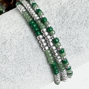 May include: Three beaded bracelets. Each bracelet features a combination of silver-coloured metal beads, dark green beads, and light green beads. The bracelets are stacked together, creating a layered effect. The background is a light grey, textured surface.