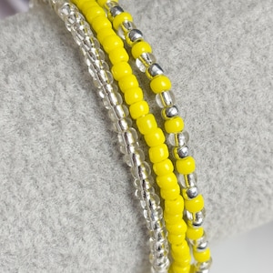 May include: Close-up of three beaded bracelets. One bracelet is made of clear beads, another of bright yellow beads, and the third combines yellow and silver beads. The bracelets are displayed on a gray surface.