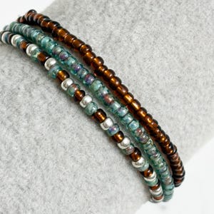 May include: Three beaded bracelets in shades of brown, teal, and silver. The bracelets are made of small, round beads strung together. The bracelets are stacked on top of each other, creating a layered look. The bracelets are displayed on a gray surface.