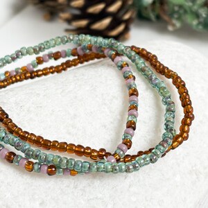 May include: Close-up of three beaded bracelets. The bracelets feature small, colorful beads in shades of brown, teal, and lavender. The bracelets are arranged in a layered style, creating a textured and visually appealing accessory.