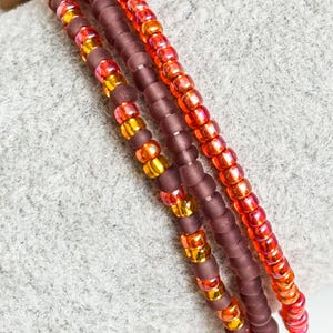 May include: Close-up of three beaded bracelets. The bracelets feature a mix of colours, including deep purple, orange, and gold. The beads are small and strung together, creating a layered effect. The bracelets are displayed on a grey surface.
