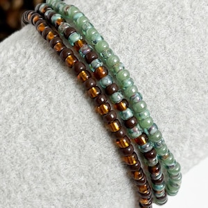 May include: Three beaded bracelets in shades of brown, amber, and green. The bracelets are made of small, round beads strung together. The beads have a glossy finish and are arranged in a linear pattern.