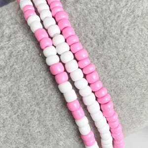 May include: Three beaded bracelets with alternating pink and white beads. The bracelets are made of small, round beads strung together. The bracelets are displayed on a gray surface.
