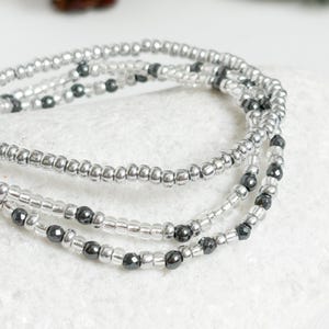 May include: Three silver beaded bracelets, each with small, clear beads and interspersed dark grey beads. The bracelets are layered and curved, resting on a white surface. The beads have a metallic sheen.