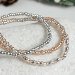 May include: Three beaded bracelets in silver and peach colours. The bracelets are made of small, round beads strung together. The bracelets are stacked, creating a layered look. The background is a light, neutral colour.