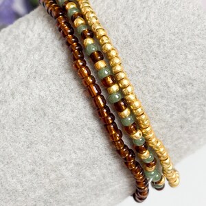 May include: A set of three beaded bracelets. One strand features translucent brown beads, another consists of metallic gold beads, and the third strand alternates between small sage green beads and metallic gold beads, all arranged in a parallel design.