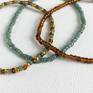 May include: Close-up of three beaded bracelets. The bracelets feature small, round beads in shades of teal, gold, and brown. The beads are strung together to form a flexible, circular shape. The bracelets are likely worn as jewellery.