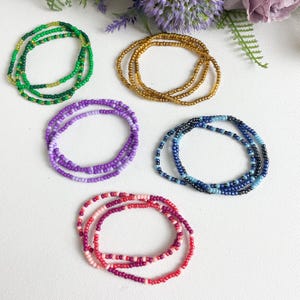 May include: Five colourful beaded bracelets, each with a different colour scheme. The bracelets are made of small, round beads and are arranged in a circle on a white surface. The colours of the bracelets are green, gold, purple, blue, and pink.