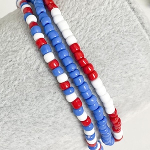 May include: Three beaded bracelets with a patriotic color scheme. Each bracelet features a repeating pattern of red, white, and blue beads. The bracelets are displayed on a light gray surface.
