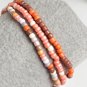 May include: Three beaded bracelets in shades of orange, pink, brown, and white. The bracelets are made of small, round beads and are arranged side by side. The bracelets are resting on a grey surface.