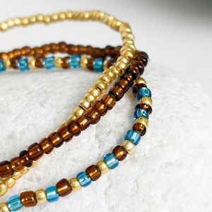 May include: A close-up shot of three beaded bracelets. One is gold, another is brown, and the third is a mix of gold, brown, and blue beads. The bracelets are stacked together. The beads are small and round.