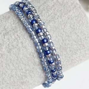 May include: Three beaded bracelets in shades of blue and silver. The bracelets are made of small, round beads strung together. The beads alternate between light blue, dark blue, and silver. The bracelets are stacked on top of each other.