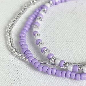 May include: Three layered beaded bracelets in a silver, white and lavender colour scheme.
