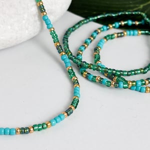 May include: A long strand of beaded jewellery featuring a repeating pattern of opaque turquoise blue, translucent emerald green, and metallic gold seed beads. The beads are strung in a consistent sequence, creating a slender, flexible, and colourful accessory.