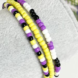 May include: A multi-strand beaded bracelet with yellow, white, purple, and black beads. The bracelet is made of small, round beads and is designed in a repeating pattern.
