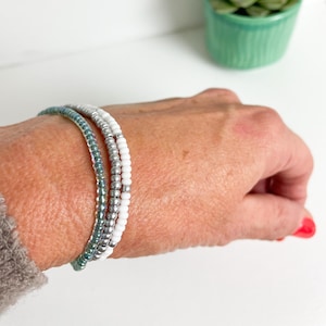 May include: A set of three beaded bracelets in shades of teal, silver, and white. The bracelets are stacked on a person's wrist.