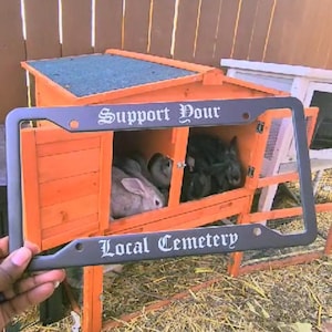 May include: A black license plate frame with the text "Support Your Local Cemetery" in white lettering. The frame is attached to a wooden rabbit hutch with several rabbits inside.