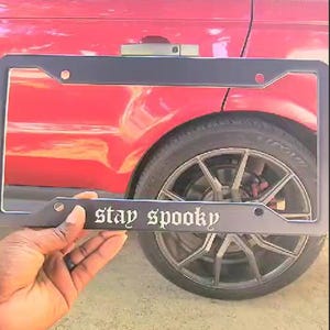 May include: Black license plate frame with the text "stay spooky" in white gothic lettering. The frame is attached to a red car.