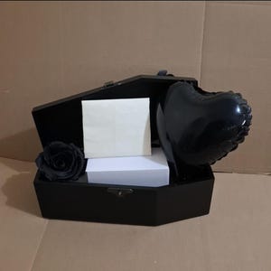 May include: A black coffin-shaped box containing a single black rose, a white rectangular envelope, a stack of white paper, and a glossy black heart-shaped balloon. The box features a metal latch on the front and is open to display the contents inside.