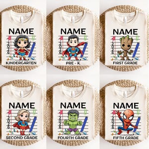 May include: Cream-colored t-shirts with superhero graphics and the word "NAME" above grade level text. The grades include Kindergarten, Pre-K, First, Second, Fourth, and Fifth Grade. Each shirt has a pencil and the year 2026.