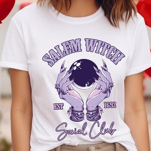 May include: White t-shirt featuring a purple graphic with the text "Salem Witch Social Club" and the year "1692." The design includes hands holding a crystal ball. The shirt is a crew neck.