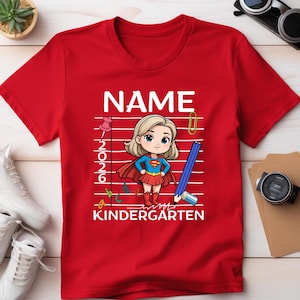May include: Red t-shirt with a cartoon illustration of a superhero girl. The shirt features the word "NAME" at the top, with "2026" and "KINDERGARTEN" below the illustration. A pencil, paperclip, and ABCs are also included.