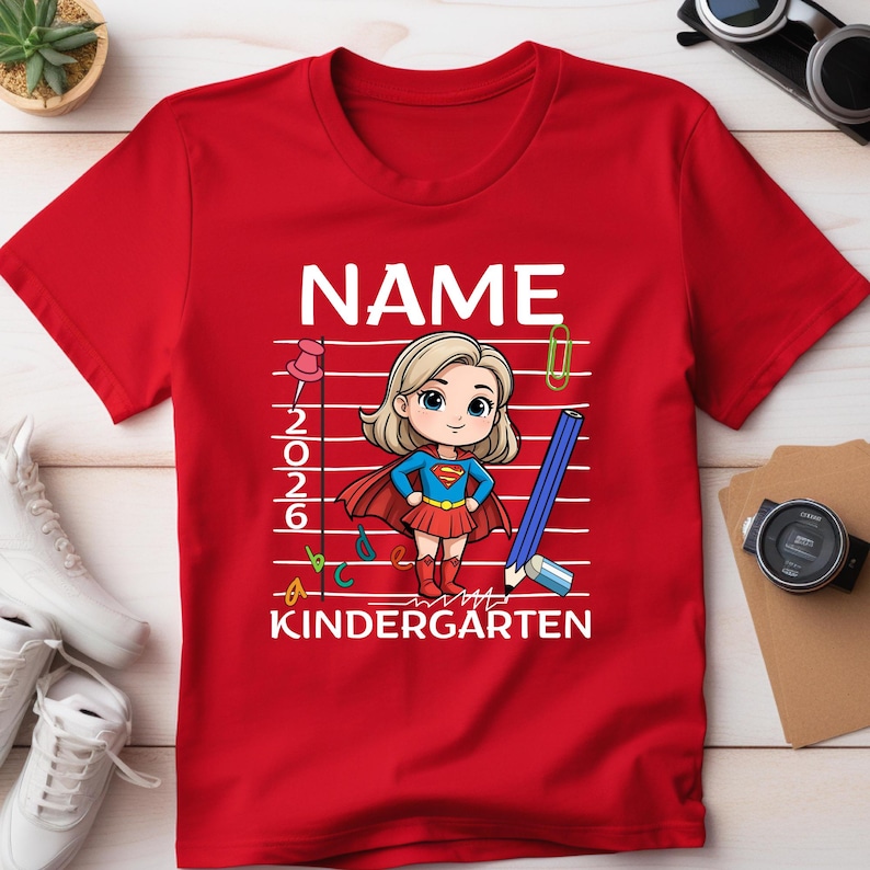 May include: Red t-shirt with a cartoon illustration of a superhero girl. The shirt features the word "NAME" at the top, with "2026" and "KINDERGARTEN" below the illustration. A pencil, paperclip, and ABCs are also included.