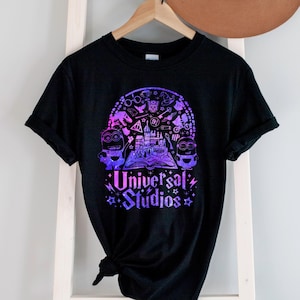 May include: Black t-shirt with a galaxy-themed graphic of popular Universal Studios attractions, including Hogwarts, the Minions, and Transformers. The text "Universal Studios" is printed in a purple and pink gradient.