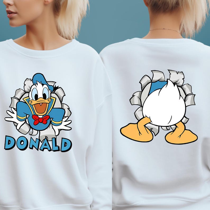 Puede incluir: Sudadera blanca con un gr&aacute;fico de dibujos animados de Pato Donald. La parte delantera muestra a Donald con un traje de marinero azul con el nombre "DONALD". La parte trasera muestra la parte trasera y los pies de Donald.