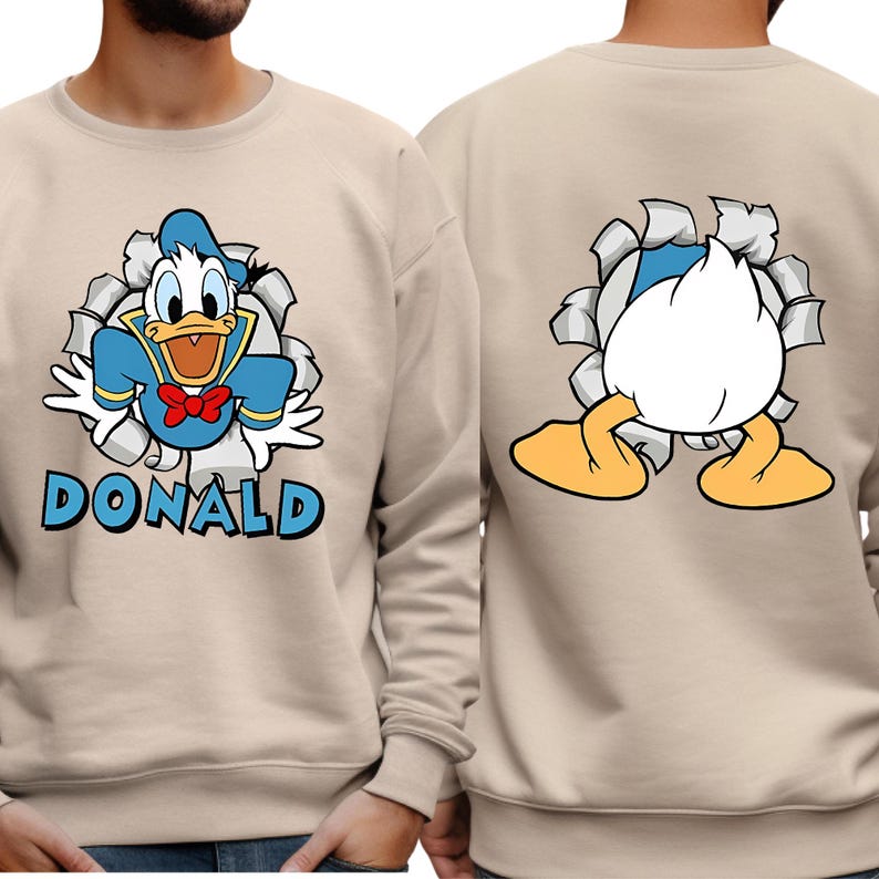 Puede incluir: Sudadera beige con un dise&ntilde;o de dibujos animados de Pato Donald. La parte delantera muestra a Donald rompiendo un fondo rasgado con el nombre "DONALD" debajo. La parte trasera muestra la parte trasera de Donald, tambi&eacute;n emergiendo de un fondo rasgado.