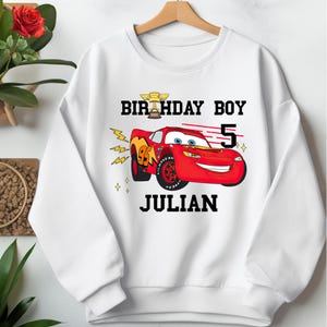 May include: White sweatshirt featuring a red cartoon race car with the text "BIRTHDAY BOY" above and "JULIAN" below. The number "5" is next to the car. The car has the number "95" on the side.