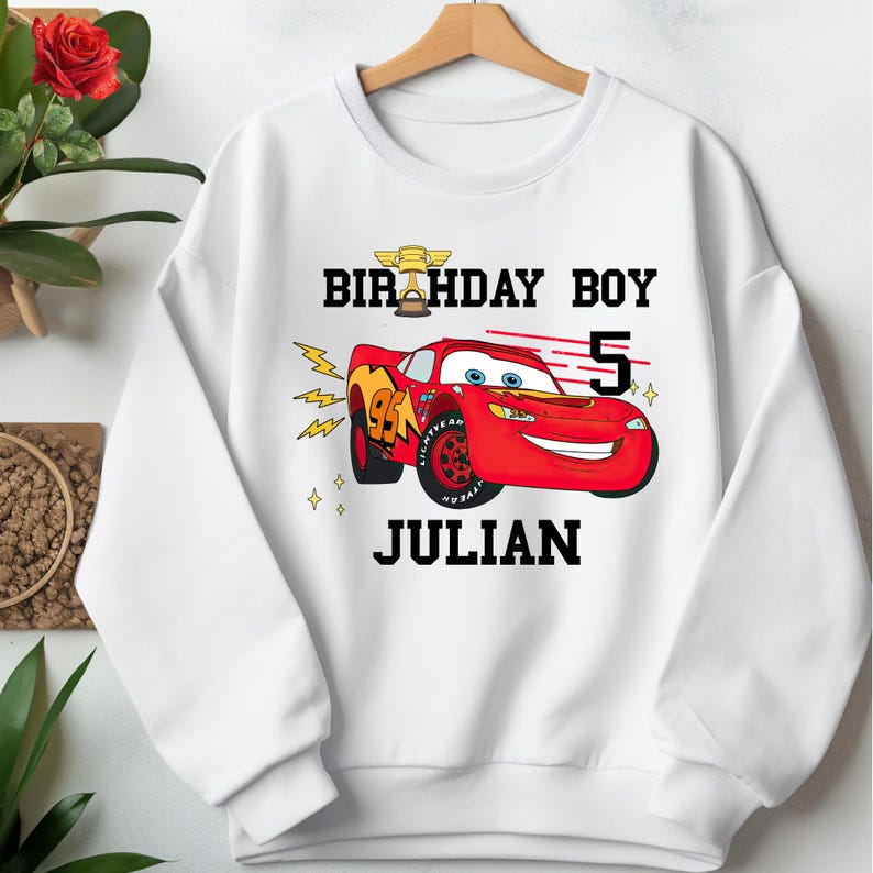 May include: White sweatshirt featuring a red cartoon race car with the text "BIRTHDAY BOY" above and "JULIAN" below. The number "5" is next to the car. The car has the number "95" on the side.