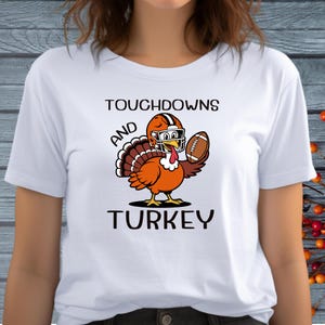 May include: A white t-shirt featuring a graphic of a cartoon turkey wearing an orange and brown football helmet and holding a brown football. The text above and below the turkey reads TOUCHDOWNS AND TURKEY in black, bold, sans-serif lettering.