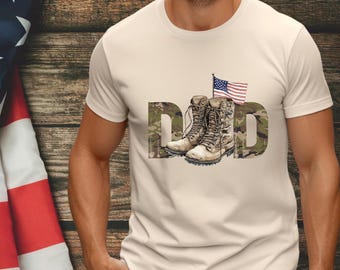 May include: Cream-colored t-shirt with a graphic design featuring the word "DAD" in a camouflage pattern, combat boots, and an American flag. The shirt is a short-sleeved crew neck.