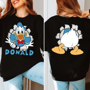 Puede incluir: Sudadera negra con un dise&ntilde;o de dibujos animados de Pato Donald rompiendo un fondo rasgado, con el nombre "DONALD" en azul. La parte trasera muestra el trasero y los pies de Pato Donald. La sudadera la lleva una persona.