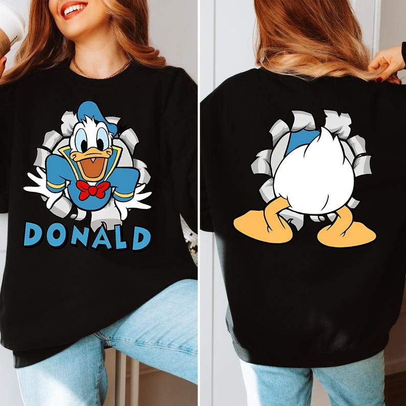 Puede incluir: Sudadera negra con un dise&ntilde;o de dibujos animados de Pato Donald rompiendo un fondo rasgado, con el nombre "DONALD" en azul. La parte trasera muestra el trasero y los pies de Pato Donald. La sudadera la lleva una persona.