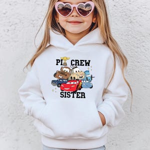 May include: White hooded sweatshirt featuring a graphic of animated cars with the text "Pit Crew Sister". The design includes characters from the movie Cars, such as Lightning McQueen and Mater. The sweatshirt has a kangaroo pocket.