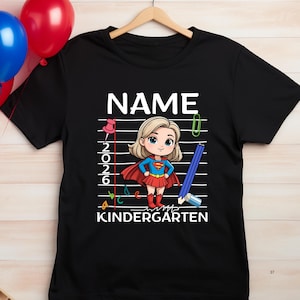 May include: Black t-shirt with a superhero graphic and the word "NAME" in white. The graphic includes a cartoon girl in a red cape and skirt, with the text "Kindergarten" below. A pencil and paperclip are also featured.