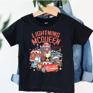 May include: Black t-shirt featuring a colorful graphic of characters from the movie Cars, including Lightning McQueen, with the text "Lightning McQueen" in red and orange. The design includes stars and a trophy.