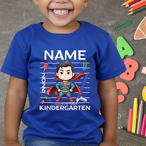 May include: A blue t-shirt featuring a cartoon Superman character with the text "NAME" above and "KINDERGARTEN" below. The design includes a pencil, pushpin, and the year "2026".