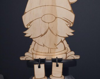 May include: A wooden gnome ornament with a red hat, a white beard, and black boots. The gnome is sitting on a black base with two clips for hanging.