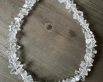 Op de afbeelding: Een ketting van heldere kwartskristallen. De ketting bestaat uit onregelmatig gevormde, gefacetteerde kwartskristallen die aan elkaar zijn geregen. De transparante kristallen reflecteren licht. De ketting wordt gepresenteerd op een houten oppervlak.