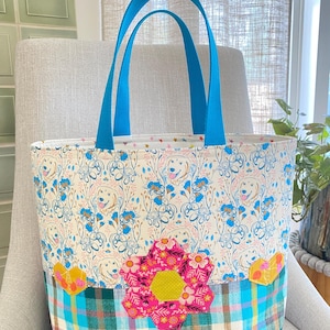 May include: A tote bag with bright blue handles featuring a top section of white fabric with a repeating golden retriever and blue floral pattern. The bottom section is a blue, green, and pink plaid fabric with a large pink floral applique.