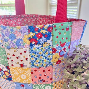 May include: A patchwork tote bag with a variety of floral patterns in blue, yellow, pink, and green. The bag has a solid pink lining and matching pink handles. The bag is designed with a quilted patchwork pattern.